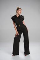 A model wearing a black wide-leg jumpsuit with dramatic ruffled shoulders and a small gold accent on the chest, posed diagonally against a light gray studio background.